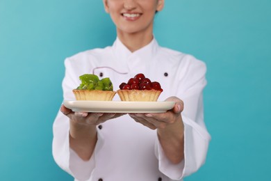 Photo of Happy professional confectioner in uniform holding plate with delicious tartlets on light blue background, closeup