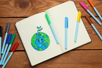 Happy Earth Day. Notebook with drawing of planet and markers on wooden table, flat lay
