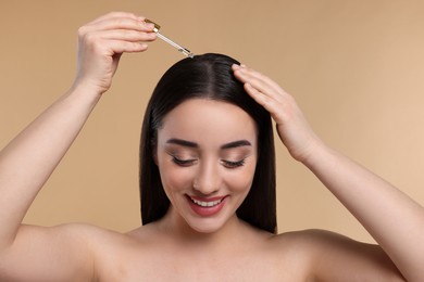 Photo of Happy young woman applying essential oil onto hair roots on beige background, closeup