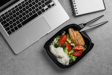 Photo of Container with tasty food, laptop, cutlery and notebook on light grey table, flat lay. Business lunch