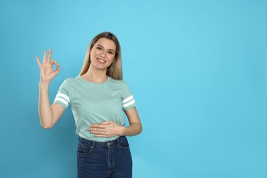 Happy woman touching her belly and showing okay gesture on light blue background, space for text. Concept of healthy stomach