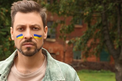 Photo of Sad man with drawings of Ukrainian flag on face outdoors. Space for text