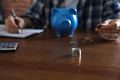 Man with piggy bank counting money at wooden table, focus on stack of coins