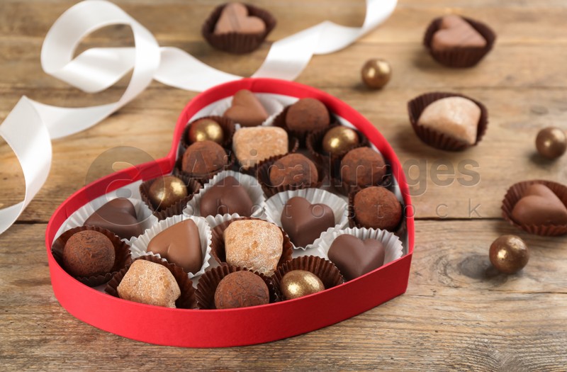 Photo of Different delicious chocolate candies in heart shaped box on wooden table