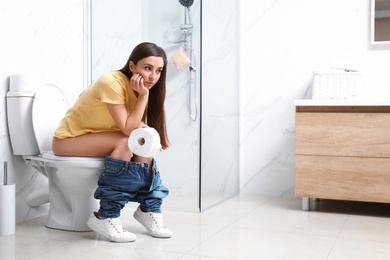 Upset woman with paper roll sitting on toilet bowl in bathroom