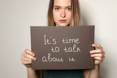 Young woman holding card with words IT'S TIME TO TALK ABOUT IT against light background