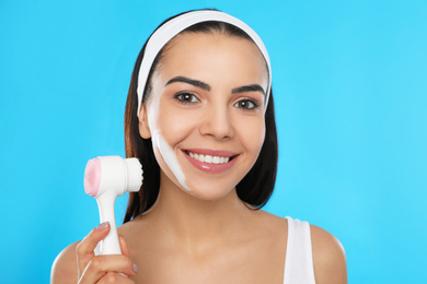 Young woman using facial cleansing brush on light blue background. Washing accessory