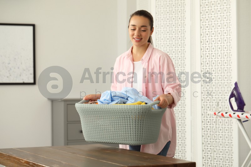 Young woman putting basket full of clean laundry on wooden table indoors