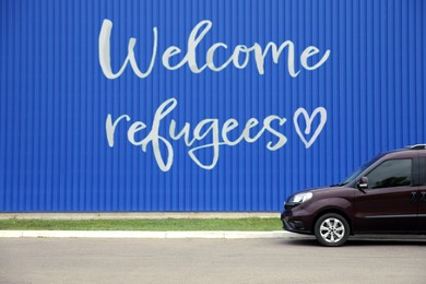 Car parked near blue wall with phrase WELCOME REFUGEES outdoors