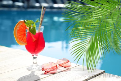 View of beautiful green tropical leaves and cocktail with sunglasses near outdoor swimming pool on sunny day