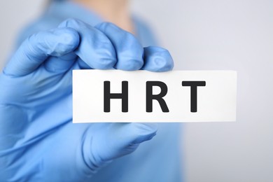 Doctor holding card with abbreviation HRT on light grey background, closeup. Hormone Replacement Therapy