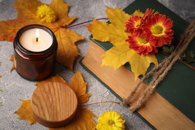 Photo of Burning scented candle, autumn leaves and book with beautiful flowers on light gray textured table