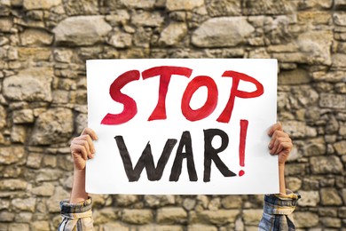 Photo of Woman holding poster with words Stop War against brick wall outdoors, closeup