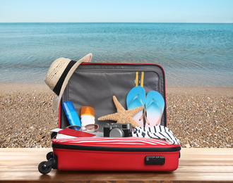 Suitcase with different beach objects on wooden table near sea