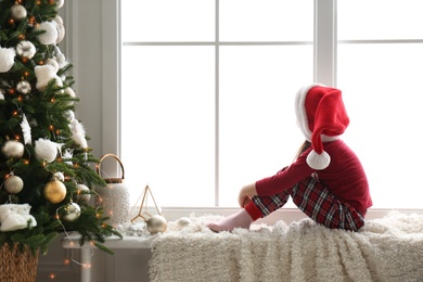 Cute little girl in Santa hat on window sill near Christmas tree at home