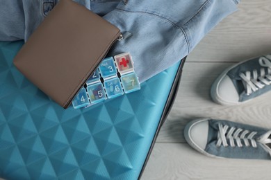 Suitcase with pill box, denim shirt and shoes on floor, flat lay