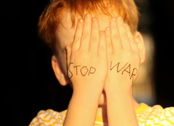 Little boy hiding face and words Stop War written on his hands outdoors, closeup