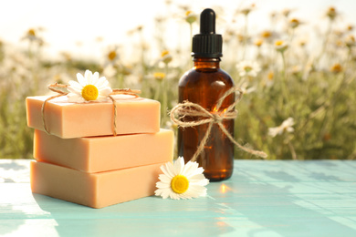 Bottle of chamomile essential oil and soap bars on light blue wooden table in field