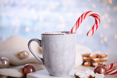 Cup of tasty cocoa with marshmallows and Christmas candy cane on white stand against blurred festive lights