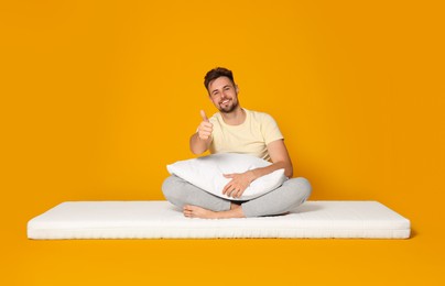 Smiling man sitting on soft mattress and showing thumb up against orange background