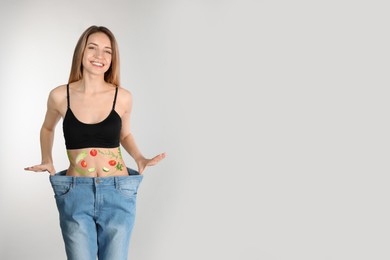 Slim woman in oversized jeans and images of vegetables on her belly against light background. Healthy eating