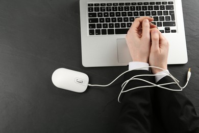 Photo of Man showing hands tied with computer mouse cable near laptop at black table, top view. Internet addiction