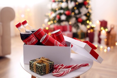 MYKOLAIV, UKRAINE - JANUARY 15, 2021: Gift box with Coca-Cola cans and bottles on table in room decorated for Christmas