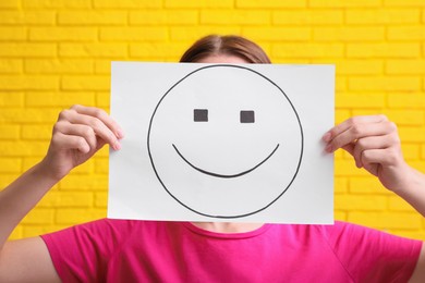 Photo of Woman hiding behind sheet of paper with happy face against yellow brick wall