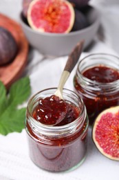 Photo of Glass jars of tasty sweet fig jam with spoon and fruits on white wooden table