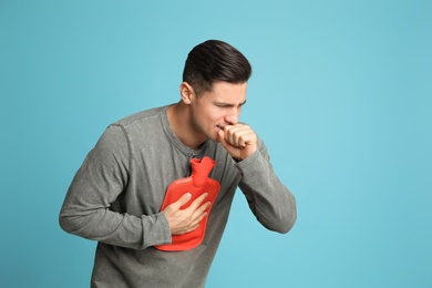 Photo of Ill man with hot water bottle coughing on light blue background