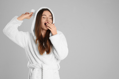 Beautiful young woman in bathrobe with funny hood  yawning on light grey background. Space for text