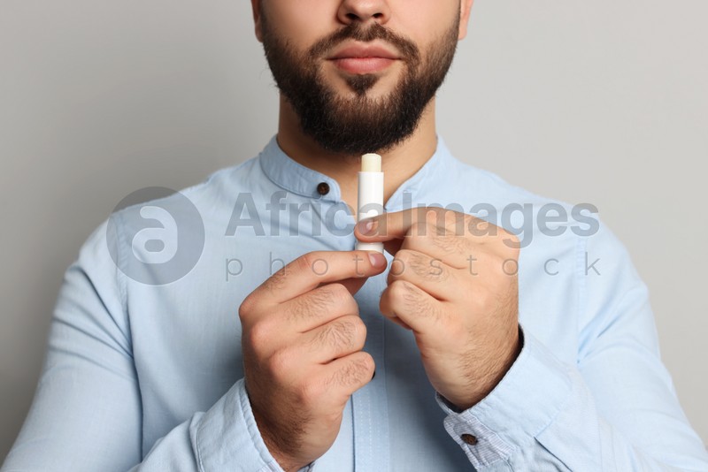 Young man applying lip balm on grey background, closeup