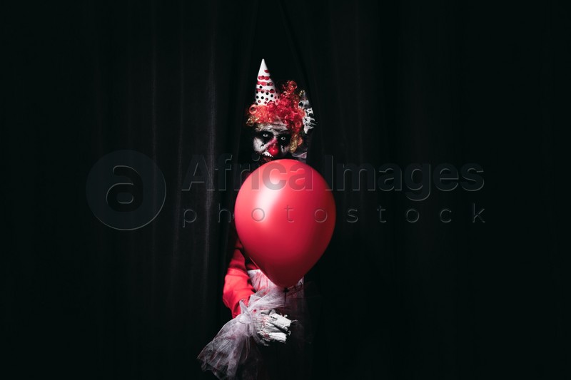 Photo of Terrifying clown with red air balloon hiding behind black curtains. Halloween party costume