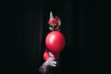 Terrifying clown with red air balloon hiding behind black curtains. Halloween party costume
