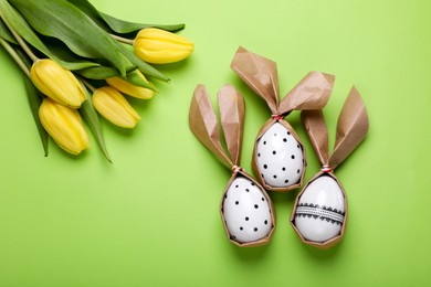 Photo of Easter bunnies made of craft paper and eggs near beautiful tulips on light green background, flat lay