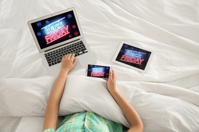 Woman using tablet, mobile phone and laptop with Black Friday announcement while lying in bed, above view