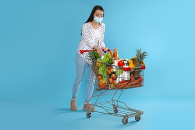 Young woman in medical mask with shopping cart full of groceries on light blue background