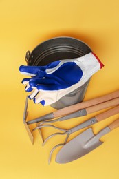 Bucket with gardening gloves and tools on yellow background, flat lay