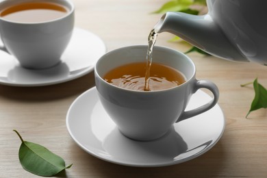 Photo of Pouring green tea into white cup with saucer and leaves on wooden table, closeup