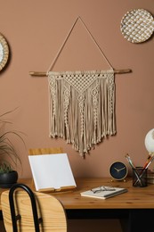Stylish macrame and wicker wall decor hanging above wooden table with stationery