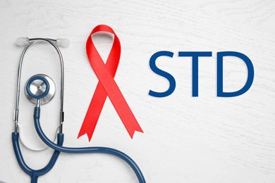 Red awareness ribbon, stethoscope and abbreviation STD on white wooden background, flat lay