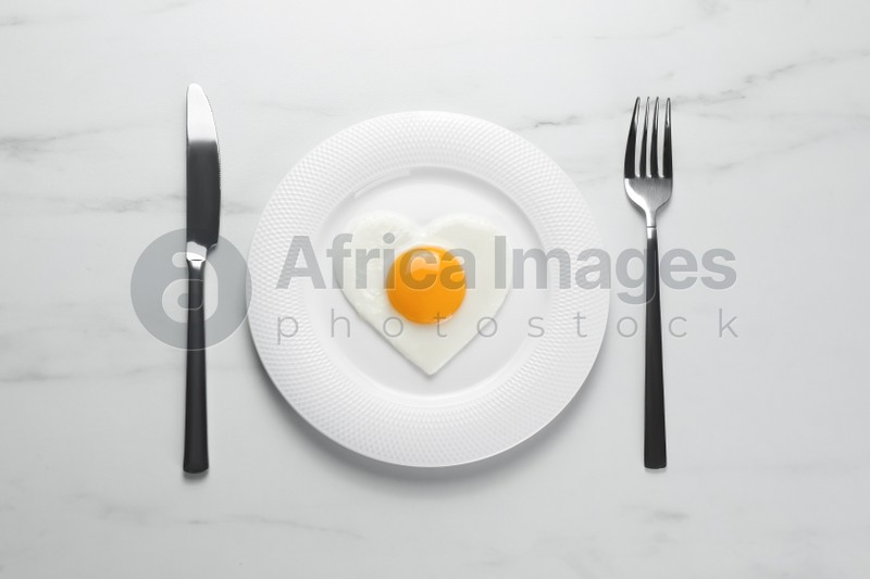 Plate with heart shaped fried egg and cutlery on white marble table, flat lay