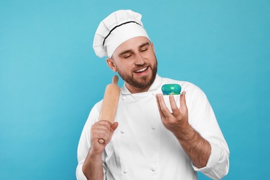 Photo of Happy professional confectioner in uniform with delicious cake and rolling pin on light blue background