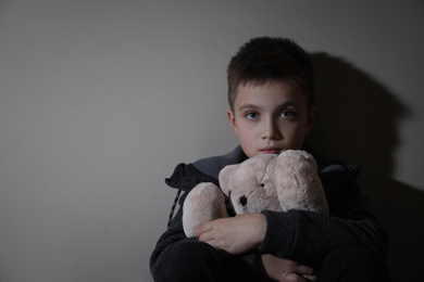 Sad little boy with teddy bear near beige wall, space for text. Domestic violence concept