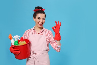 Young housewife holding bucket with cleaning supplies on light blue background, space for text