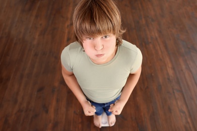 Emotional overweight boy standing on floor scales indoors, above view