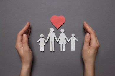 Photo of Woman protecting paper family figures and red heart on grey background, top view. Insurance concept