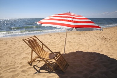Deck chair near red and white striped beach umbrella on sandy seashore