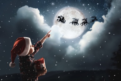 Cute little boy looking at Santa Claus with reindeers in sky on full moon night. Christmas holiday