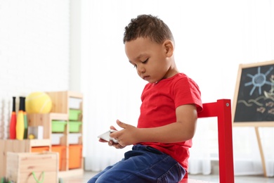 Cute little African-American child playing on mobile phone in kindergarten, space for text. Indoor activity